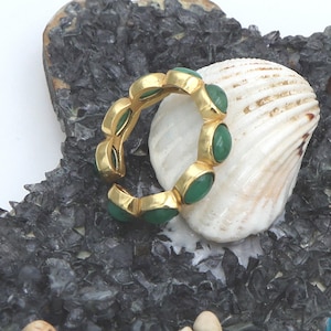 May include: A gold-colored ring with oval-shaped green stones. The ring is set against a backdrop of a seashell and dark, textured stones, creating a contrast of colors and textures. The ring's design is simple yet elegant.