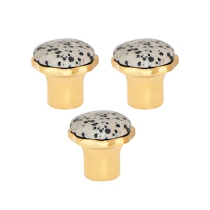 Op de afbeelding: Drie kastknoppen met een goudkleurige basis en een gespikkelde, gebroken witte en zwarte stenen bovenkant. De knoppen hebben een afgeronde vorm en staan tegen een witte achtergrond. Deze decoratieve knoppen zijn geschikt voor meubels en woondecoratie.