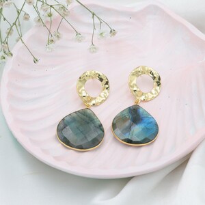 May include: A pair of gold earrings with a hammered texture, each featuring a teardrop-shaped, iridescent, blue-green gemstone. The earrings are displayed on a pink, scalloped tray, with white flowers in the background. Jewellery.