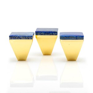 Può includere: Tre anelli dorati dal design geometrico. Ogni anello presenta una pietra quadrata blu scuro incastonata su una fascia dorata. Gli anelli sono esposti su una superficie bianca, con i loro riflessi visibili sotto.