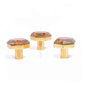 Op de afbeelding: Drie decoratieve kastknoppen. Elke knop heeft een facetgeslepen, juweelachtige bovenkant met oranje, groene en bruine patronen, geplaatst op een goudkleurige basis. De knoppen zijn ontworpen voor meubels en woondecoratie.