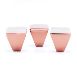 Pode incluir: Três bancos modernos e geométricos. Cada banco tem uma base em forma de diamante em ouro rosa e um tampo retangular branco. Os bancos estão dispostos em fila contra um fundo branco, mostrando seu design contemporâneo e acabamento metálico.