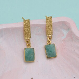 May include: A pair of gold-colored earrings with rectangular, light green gemstones. The earrings have a textured, rectangular top piece and a small chain connecting to the gemstone. The earrings are displayed on a pink and white tray.