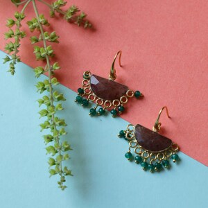 May include: A pair of gold-colored earrings with a half-moon shaped, maroon-colored stone. The earrings have a series of small green beads and a gold-colored hook. The background is a mix of pink and blue.