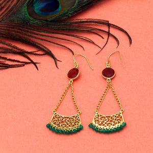 May include: A pair of gold-colored earrings with a red gemstone at the top and a decorative filigree design. The earrings have a chain and small green beads at the bottom. A peacock feather is in the background.