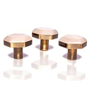 Pode incluir: Três puxadores decorativos para armários com design geométrico. Cada puxador apresenta um topo octogonal creme com uma borda marrom escura e uma base dourada. Os puxadores estão sobre um fundo branco.