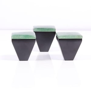 May include: Three contemporary stools with a geometric design. Each stool features a black, trapezoidal base and a square, light green cushioned top. The stools are arranged in a row against a white backdrop.