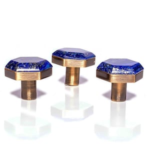 May include: Three cabinet knobs with a geometric design. Each knob features a dark blue, octagonal gemstone top with gold-coloured metal trim and a cylindrical brass stem. The knobs are set against a white background.