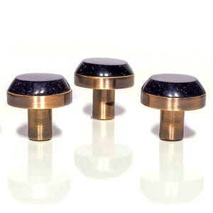 May include: Three cabinet knobs with a dark blue, speckled top, a gold-coloured band, and a brass-coloured stem. The knobs are displayed against a white background. Each knob is approximately 3 cm in diameter.