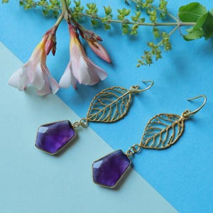 Könnte beinhalten: Ein Paar goldfarbene Ohrringe mit Blattdesign und einem facettierten, tiefvioletten Edelstein. Die Ohrringe hängen vor einem blauen Hintergrund mit rosa und weißen Blüten.