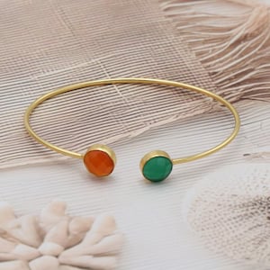 Könnte beinhalten: Ein offenes Armreif-Armband in Goldoptik mit einem runden, orangefarbenen Edelstein an einem Ende und einem runden, grünen Edelstein am anderen Ende. Das Armband befindet sich vor einem neutralen Hintergrund mit einer Feder.