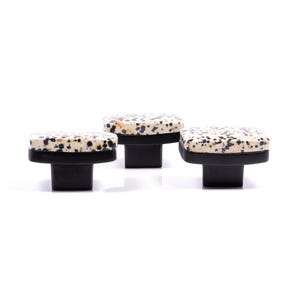 May include: Three cabinet knobs with a speckled, off-white and black stone top and a black base. The knobs are square and have a modern design. The knobs are suitable for drawers and cabinets.