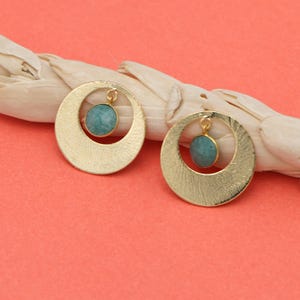 May include: A pair of gold-colored, textured hoop earrings with a small, round, turquoise-colored stone suspended in the center of each hoop. The earrings are set against a coral background with a piece of driftwood.