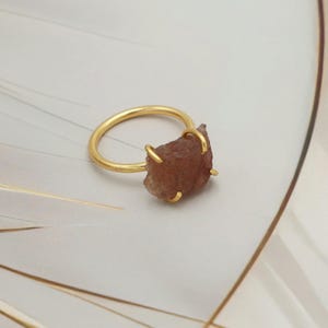 Pode incluir: Um anel dourado com uma gema bruta castanho-avermelhada. A gema é mantida no lugar por quatro garras. A banda do anel é simples e lisa. O anel é exibido sobre um fundo neutro.