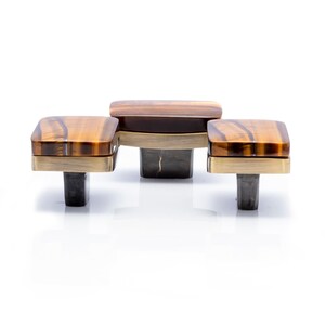 May include: Three-piece cabinet hardware set. Each piece features a square, tiger-eye-patterned top with a brass-colored border. The central piece has a wooden top. The hardware is mounted on dark gray, rectangular posts.