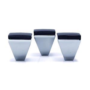 Könnte beinhalten: Drei geometrische, moderne Schrankknöpfe. Jeder Knopf hat eine dreieckige, silberfarbene Basis, die von einem rechteckigen, schwarzen Akzent gekrönt wird. Die Knöpfe sind in einer Reihe vor einem weißen Hintergrund angeordnet.