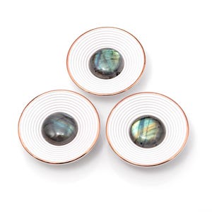 Puede incluir: Tres posavasos decorativos blancos y cobrizos. Cada posavasos presenta una piedra central ovalada e iridiscente, engastada en un diseño blanco de círculos concéntricos. Los posavasos están dispuestos sobre un fondo blanco.