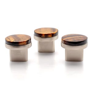 May include: Three cabinet knobs with a natural tiger eye stone top and a brushed nickel base. The knobs have a round top and a rectangular base. The tiger eye stone has brown and gold tones.