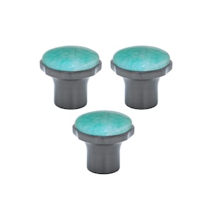 Może przedstawiać: Trzy dekoracyjne gałki do szafek. Każda gałka ma okrągłą, turkusową górę o teksturowanym wyglądzie, osadzoną na ciemnoszarej, cylindrycznej podstawie. Gałki są przeznaczone do mebli lub szafek.