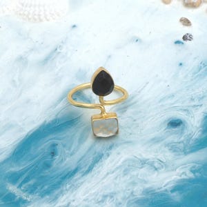 May include: A gold-colored ring featuring a black teardrop-shaped stone and a square, white stone. The ring has a unique, open design with the stones set on opposite ends of the band. The ring is displayed on a blue and white marbled surface.