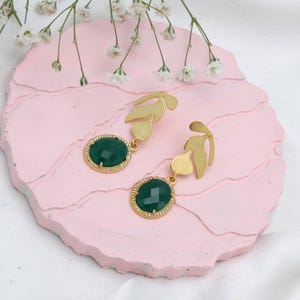 May include: Gold-coloured leaf earrings featuring oval, faceted, emerald-green gemstones. The gemstones are set in a gold frame with small, clear crystals. The earrings are displayed on a pink, textured surface with white flowers.