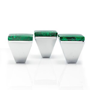 Può includere: Tre anelli argentati con top quadrati verde malachite. Gli anelli hanno una forma geometrica trapezoidale. La malachite ha un motivo a vortice. Gli anelli sono esposti su una superficie bianca.