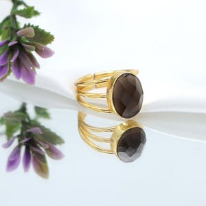 May include: A gold-toned ring with a large, faceted, oval-shaped smoky quartz gemstone. The ring has a triple-band design and is displayed on a reflective surface. The gemstone is a dark, translucent brown color.