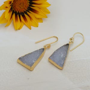 May include: Gold-coloured earrings featuring a triangular, pale blue stone with gold edging. The earrings have a hook design. A yellow flower with brown markings is in the background.
