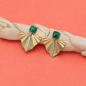 May include: A pair of gold-colored earrings with a fan-like design. Each earring features a square, emerald-green stone at the top. The earrings are set against a peach-colored background and a white, woven element.