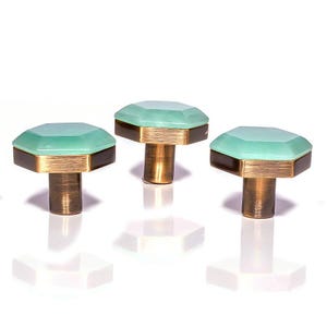 May include: Three decorative cabinet knobs with a geometric design. Each knob features a light green, faceted top, a gold-coloured band, and a dark brown base. The knobs are set against a white background.