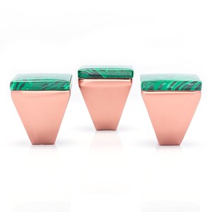 Op de afbeelding: Drie decoratieve kastknoppen. Elke knop heeft een vierkante, groene malachiet bovenkant en een koperkleurige, geometrische basis. De knoppen zijn gerangschikt op een witte achtergrond, wat hun ontwerp en kleurcontrast benadrukt.