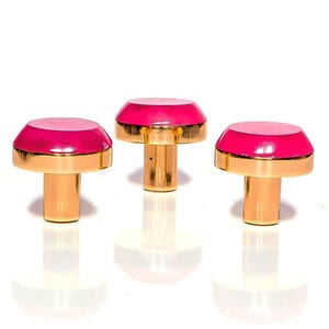 May include: Three mushroom-shaped cabinet knobs with gold stems and two-tone pink tops. The tops have a lighter pink centre and a darker pink border. The knobs are displayed on a white surface. These knobs are suitable for furniture.