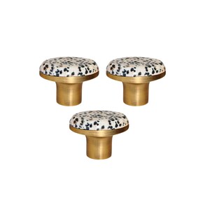 Könnte beinhalten: Drei dekorative Schrankknöpfe mit goldfarbenem Sockel und rundem Oberteil. Die Oberseite ist mit einem cremefarbenen Stoff mit schwarzen Sprenkeln bedeckt. Diese Knöpfe sind für Möbel und Heimdekoration konzipiert.