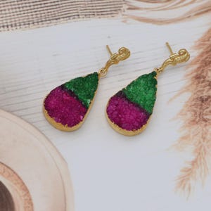 May include: A pair of gold-coloured earrings with a teardrop design. Each earring features a section of green and magenta-coloured, textured stone. The earrings have a decorative, curved top.