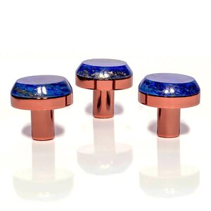 May include: Three decorative cabinet knobs. Each knob has a copper-coloured base and stem, topped with a blue and white design with gold flecks. The knobs are arranged in a row against a white background.