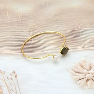May include: A gold-colored open cuff bracelet featuring a square, dark gemstone and a small, round white pearl. The bracelet has a delicate, minimalist design, perfect for everyday wear. The bracelet is displayed on a light-colored surface.