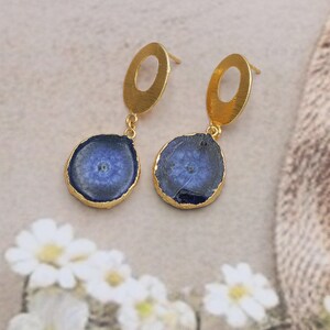Pode incluir: Um par de brincos dourados com pingentes de pedra azul circular. As pedras têm bordas douradas e padrões únicos. Os brincos têm fecho de pressão e parte superior oval. As joias são exibidas em um fundo neutro.