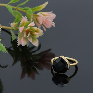 Puede incluir: Un anillo dorado con una piedra preciosa negra en forma de pera. El anillo está sobre un fondo oscuro con un arreglo floral de flores rosas y verdes. La piedra preciosa está facetada, reflejando la luz.