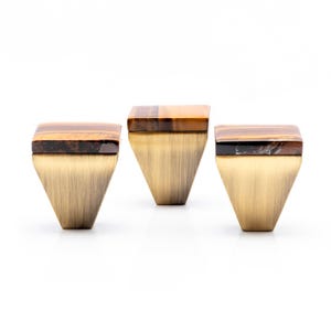 May include: Three cabinet knobs with a trapezoid design. Each knob has a brass-coloured base and a rectangular top made of tiger's eye stone. The tiger's eye stone features brown and gold tones.