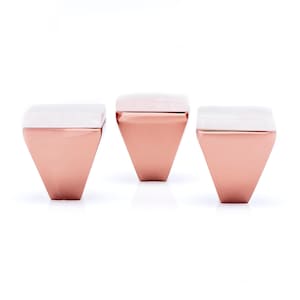 Pode incluir: Três bancos modernos e geométricos. Cada banco apresenta uma base em forma de diamante em ouro rosa e um tampo retangular branco. Os bancos estão dispostos em fila sobre um fundo branco, mostrando seu design minimalista e acabamento metálico.