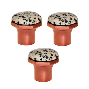 May include: Three cabinet knobs with a speckled stone top and copper-coloured base. The stone is off-white with black spots. The base has a hexagonal top and a cylindrical stem. The knobs are suitable for cabinets.