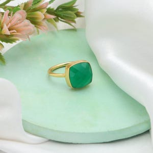 Pode incluir: Um anel de ouro com uma grande pedra preciosa verde facetada de corte quadrado. O anel tem um design simples e elegante com uma banda e engaste de ouro. A pedra preciosa é de um verde vibrante, sobre uma superfície verde claro.