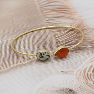 May include: A gold-coloured bangle bracelet with two gemstone features. One is a speckled, oval-shaped stone, and the other is a teardrop-shaped, orange-red stone. The bracelet is set against a neutral, textured background.