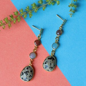 Może przedstawiać: Para złotych kolczyków wiszących. Każdy kolczyk ma duży, nakrapiany kamień w kształcie owalu na dole, z trzema mniejszymi, kolorowymi kamieniami powyżej. Kolczyki są umieszczone na różowym i niebieskim tle.