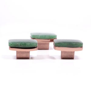Op de afbeelding: Drie decoratieve kastknoppen met een rechthoekige groene stenen bovenkant en een koperkleurige basis. De knoppen worden weergegeven tegen een witte achtergrond, die hun ontwerp en kleurcontrast benadrukt. Deze knoppen zijn ideaal om een vleugje elegantie aan meubels toe te voegen.