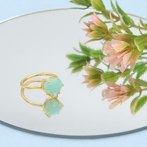 Pode incluir: Um anel dourado com uma pedra preciosa verde clara. O anel está sobre uma superfície refletora, com um arranjo floral ao fundo. A pedra preciosa tem um formato irregular único.