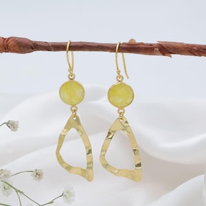 May include: A pair of gold-colored dangle earrings. Each earring features a round, pale yellow stone at the top, connected to a hammered, tear-drop shaped pendant. The earrings hang from a gold-colored hook. The background is a white, draped fabric.