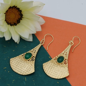 May include: Gold-coloured earrings with a fan-shaped design. Each earring has a green oval stone and detailed filigree. The earrings have a textured surface and a hook closure. A white flower with a yellow centre is in the background.