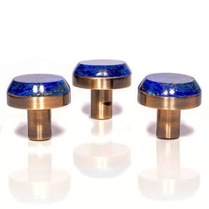 Puede incluir: Tres pomos decorativos para armarios. Cada pomo presenta una base de latón y una parte superior moteada en azul y dorado. Los pomos están dispuestos en fila sobre un fondo blanco, con sus reflejos visibles debajo.