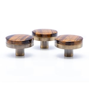 May include: Three cabinet knobs with a round, tiger-eye-patterned top and a brass-coloured base. The knobs are set against a white background, highlighting their design. These knobs are ideal for adding a touch of elegance to furniture, with a diameter of approximately 4 cm.